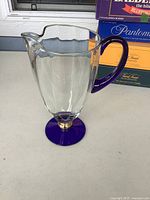 Full side view of clear glass pitcher showing cobalt blue handle and base with gold trim