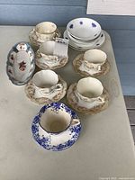 All pieces grouped on table: four cream floral teacup sets, Royal Albert Dainty Blue set, stack of violet-sprig bowls and plates, oval floral dish