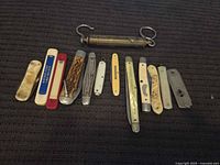 Group shot of twelve pocket knives lined below a brass tubular balance scale
