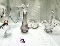 Group view of all five glass items: oil lamp, rectangular vase, smokey decanter with bird stopper, small clear basket, large etched bride’s basket