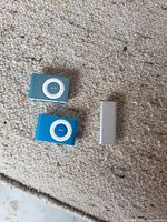 Top view of two 2nd-gen blue/teal iPod Shuffles and one 3rd-gen silver stick model on carpet