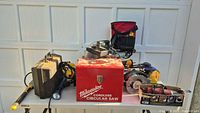 Overall view of lot on table showing Milwaukee box, miter saw, nailers, router, circular saw, flashlight, tool cases and accessories