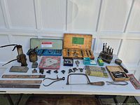 Overview of entire lot on table showing oil cans, shaper cutter box, drill bits, hacksaw, rulers