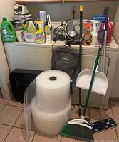 Lot overview showing bubble wrap rolls, cleaning products on appliance top, broom, dustpan, mop, fans, backpack