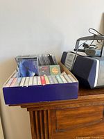Box of assorted audio cassettes alongside Sony boombox on wooden surface