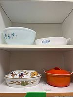 Shelf view showing snowflake mixing bowl, CorningWare Cornflower dish, fruit motif lidded casserole, and orange Le Creuset Dutch oven