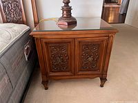 Front angled view of one nightstand showing carved doors, glass top, feet