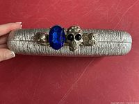 Side view of silver textured clutch showing knuckle handle with blue stone and skull embellishments