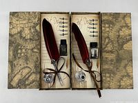 Two opened presentation boxes each holding red feather quill pen, ink bottle, metal stand and nibs