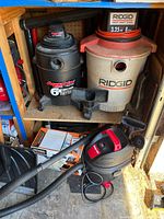 Shelf with Ridgid beige/orange 8 gal vac, Aqua Vac black 6 gal vac, small gray/red vac, hoses and boxes