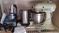 All three appliances on shelf: Ninja food processor base with bowl, KRUPS stainless kettle, KitchenAid mixer with bowl