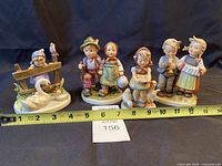 Front view of four Hummel figurines with measuring tape indicating size