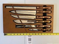 Full view of six knives in brown tray with measuring tape for scale
