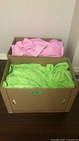 Two cardboard boxes on floor, top containing pink linens, bottom containing green linens