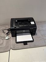 Front view of HP LaserJet P1102 printer with paper in output tray and cords beside