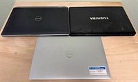 Three closed laptops showing lids: two Dell (black, silver) and one Toshiba (black with logo)