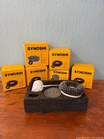Overall lot showing scrubber body in foam, grey brush head, black pad head, and five yellow Synoshi boxes