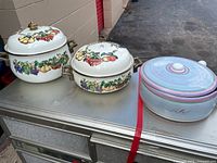 Three cookware pieces on metal surface: two Kensington Garden fruit motif pots with lids, one pastel swirl casserole with lid