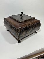 Three-quarter view of footed jewelry box showing floral embossing, nailhead trim, and finial