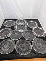 All 11 glass plates arranged: top row grapevine border saucers, bottom rows Fleur pattern plates