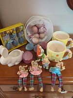 All items arranged on dresser: mugs, planter, bunny figurines, cloth ornaments, boxed and loose eggs
