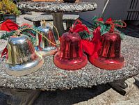 Group of six metallic-finish plastic Christmas bells on outdoor table, showing three red and three silver units with bows and greenery