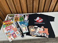 Overview of all items on table: T-shirt, magazines, comic, Beanie Babies, belt, card packs, videocassette, postcards