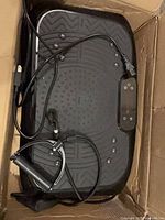 Top view of vibration plate in box with resistance bands and power cord visible