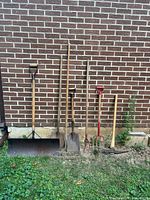 Five long-handled garden tools leaning against brick wall