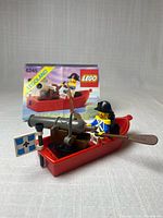 Set assembled: red boat with cannon, oars, flag, minifigure; instruction booklet behind
