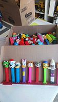 Open cardboard box filled with loose PEZ dispensers; front row shows 8 different character designs