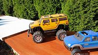 Yellow Hummer-style RC truck next to blue pickup RC truck on wooden surface outdoors