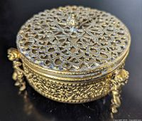 Closed brass trinket box showing pierced lid and cherub side supports