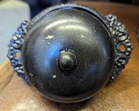 Front view of black metal Victorian doorbell showing central bell dome and side scroll mounts