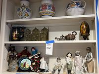 Overview of three shelves showing ceramic vases, jar, tureen, pitcher, metal panel, teapot, covered dish, icon, dolls, tin and multiple porcelain figurines