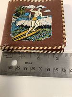 Front of brown wallet showing surfer graphic and whipstitch trim with ruler for scale