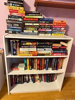 Front view of white bookcase filled with three levels of paperback novels; additional stacks on top