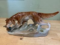 Left/front view of Royal Dux glazed porcelain figurine; shows dog retrieving duck and maker’s gold foil label on base