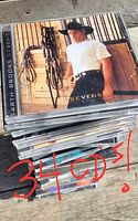 Stack of jewel-case CDs, top case Garth Brooks “Sevens”, handwritten note ‘34 CDs!’