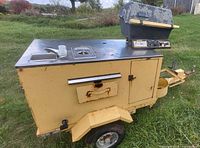 Left-front view of yellow steel hot dog cart trailer showing sink, grill, storage cabinet, wheels, round holders