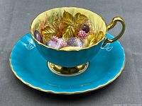 Tea cup resting on saucer showing turquoise exterior, hand-painted fruit interior, gilt detailing