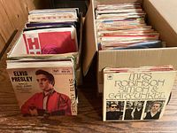 Two rows of 45 RPM records in sleeves within cardboard box; Elvis Presley Strictly Elvis EP and Simon & Garfunkel Mrs. Robinson single at front