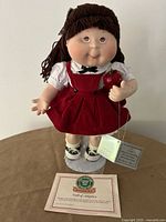 Front view of doll on stand holding apple with certificate displayed