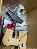 Box of Dyson vacuum parts showing wall dock, charger, combination nozzle, sealed plastic-bag items