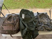 Three bags on table: multicam backpack, green duffel, digital camo backpack