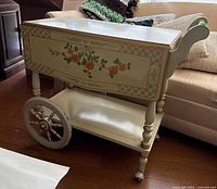 Side view of painted folding tea cart showing hinged leaves, floral motif, large wheel, lower shelf