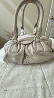 Front view of cream pebbled leather satchel with rolled handles and pleated panels