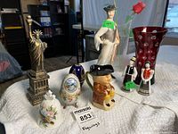 Group shot showing all items on towel: porcelain figurines, metal Statue of Liberty, decorative eggs, bell, glass vase with rose