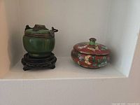 Both decorative vessels side by side within a niche: green stone lidded jar on carved stand at left, red floral cloisonné covered box at right