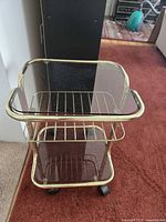Front angled view showing gold metal frame, glass shelves, central rack shelf and caster wheels